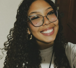 Foto da integrante Ana Clara, ela usa óculos, tem cabelo encaracolado solto e está sorrindo.