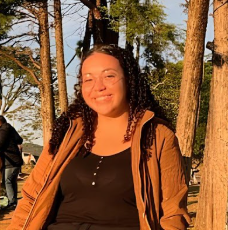Foto da integrante do projeto Ana Carolina. Ela tem cabelo encaracolado semi preso, castanho escuro,  usa óculos, está ao ar livre usando uma blusa de frio marrom.